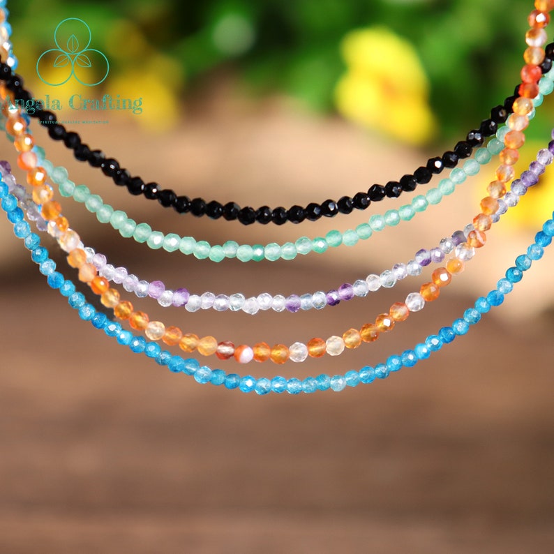 May include: A collection of delicate beaded necklaces in various colors, including black, green, purple, orange, and blue. Each necklace is composed of small, faceted beads, creating a sparkling effect. The necklaces are displayed against a blurred background.