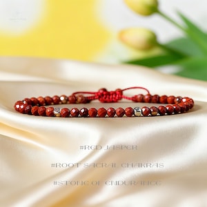 May include: Red jasper beaded bracelet with silver accents. The bracelet is adjustable with a red cord and features the text "#RED JASPER", "#ROOT SACRAL CHAKRAS", and "#STONE OF ENDURANCE".