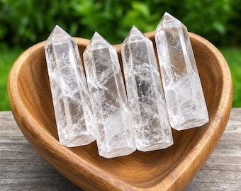 May include: Four clear, faceted quartz crystal points arranged upright inside a smooth, heart-shaped wooden bowl. Each crystal features a hexagonal shape with a pointed top, displaying natural internal fractures and transparent, colorless surfaces.