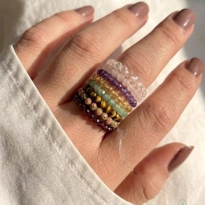 May include: A stack of seven beaded rings worn on a finger. The rings feature small, faceted, round gemstone beads in various colors including clear, light pink, purple, yellow, light green, brown, and dark red, arranged in a vertical sequence.