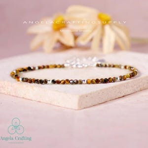 May include: A delicate bracelet featuring small, faceted tiger's eye beads in shades of brown and gold, interspersed with silver-colored beads. The bracelet is displayed on a light pink surface, with the text "ANGELACRAFTINGSUPPLY" and "Minimalist Jewelry" visible.