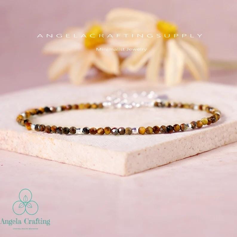 May include: A delicate bracelet featuring small, faceted tiger's eye beads in shades of brown and gold, interspersed with silver-colored beads. The bracelet is displayed on a light pink surface, with the text "ANGELACRAFTINGSUPPLY" and "Minimalist Jewelry" visible.