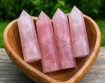 May include: Four polished rose quartz crystal points with a hexagonal shape and pointed tips, arranged upright inside a smooth, heart-shaped wooden bowl. The crystals are translucent, pale pink, and feature natural internal fractures and inclusions.