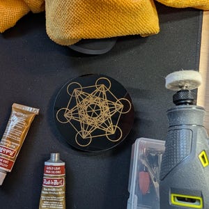 May include: A circular black coaster featuring a gold-colored geometric Metatron's Cube design. Included are two tubes of Rub 'n Buff wax metallic finish in gold leaf and antique gold, a rotary tool with a felt polishing bit, and a yellow cloth.