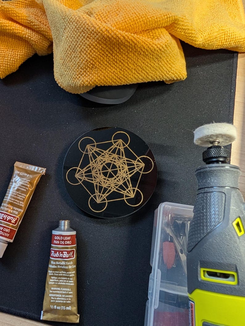 May include: A circular black coaster featuring a gold-colored geometric Metatron's Cube design. Included are two tubes of Rub 'n Buff wax metallic finish in gold leaf and antique gold, a rotary tool with a felt polishing bit, and a yellow cloth.