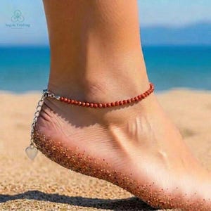 Puede incluir: Una tobillera de cuentas rojas con una cadena plateada y un dije de corazón. La tobillera se usa en un pie en una playa de arena. La tobillera está hecha de pequeñas cuentas redondas.