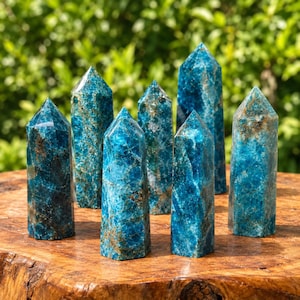 Op de afbeelding: Een verzameling van zes blauwe apatiet kristaltorens met puntige toppen. Elke steen heeft een diepblauwe kleur met natuurlijke bruine en witte mineraalinsluitingen, een gepolijst, gefacetteerd oppervlak en een zeshoekige kolomvorm.
