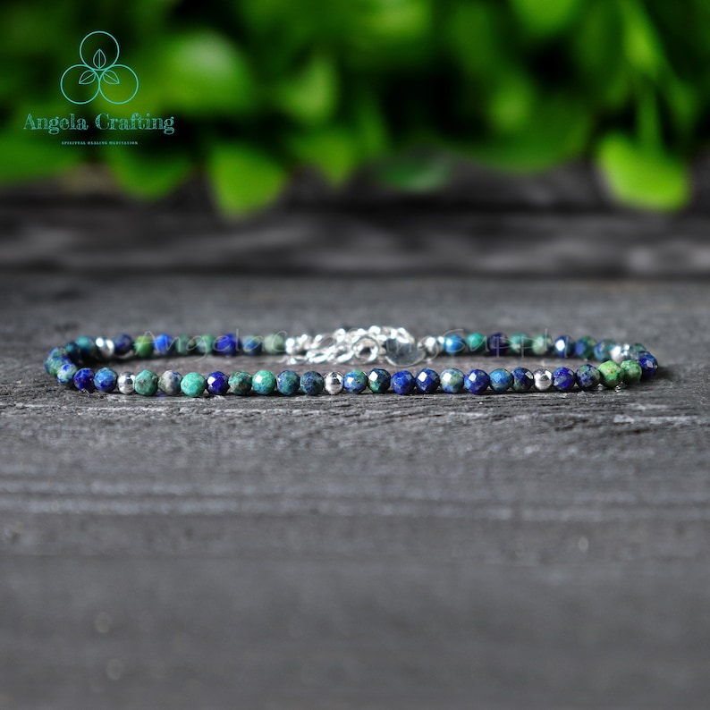 May include: A delicate bracelet made with small, faceted blue and green gemstone beads and silver spacer beads. The bracelet is on a wooden surface.