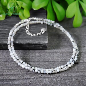 May include: A delicate necklace featuring small, round, white and gray beads strung together in a double strand. The necklace has a silver-colored chain and clasp. The background is a dark gray surface with green foliage.