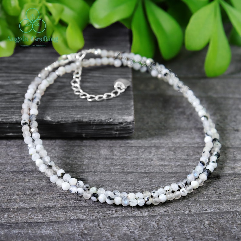 May include: A delicate necklace featuring small, round, white and gray beads strung together in a double strand. The necklace has a silver-colored chain and clasp. The background is a dark gray surface with green foliage.