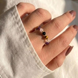 May include: A beaded ring featuring small, round, faceted gemstones in shades of purple, light pink, black, and golden brown, strung together on a thin, flexible band. The ring is worn on a finger, showcasing the alternating colors and textures.