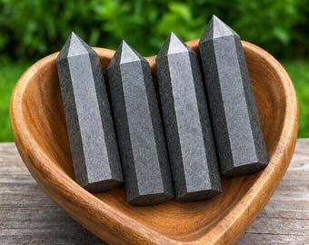 Può includere: Quattro punte in pietra grigio scuro di forma esagonale con estremità appuntite, disposte verticalmente all'interno di una ciotola in legno a forma di cuore. Le colonne in pietra presentano una superficie opaca e ruvida nel contenitore.