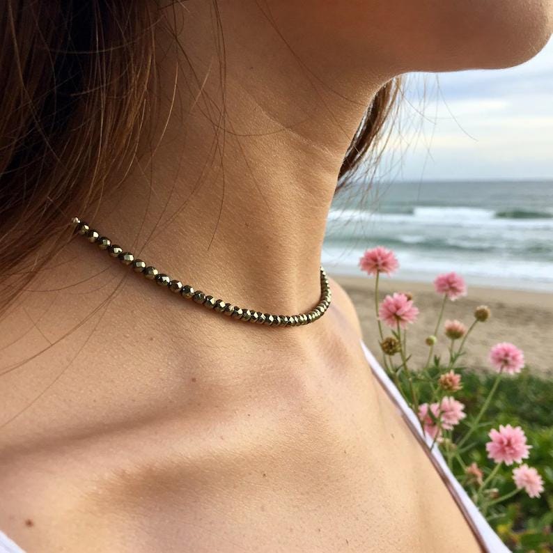 May include: A delicate necklace made of small, faceted, gold-coloured beads. The necklace is worn around the neck. The background shows a beach and ocean. Pink flowers are in the foreground.