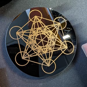 May include: A circular black coaster featuring a gold-colored Metatron's Cube geometric design etched onto the surface. The design consists of interconnected circles and straight lines forming a complex, symmetrical pattern centered on the disc.