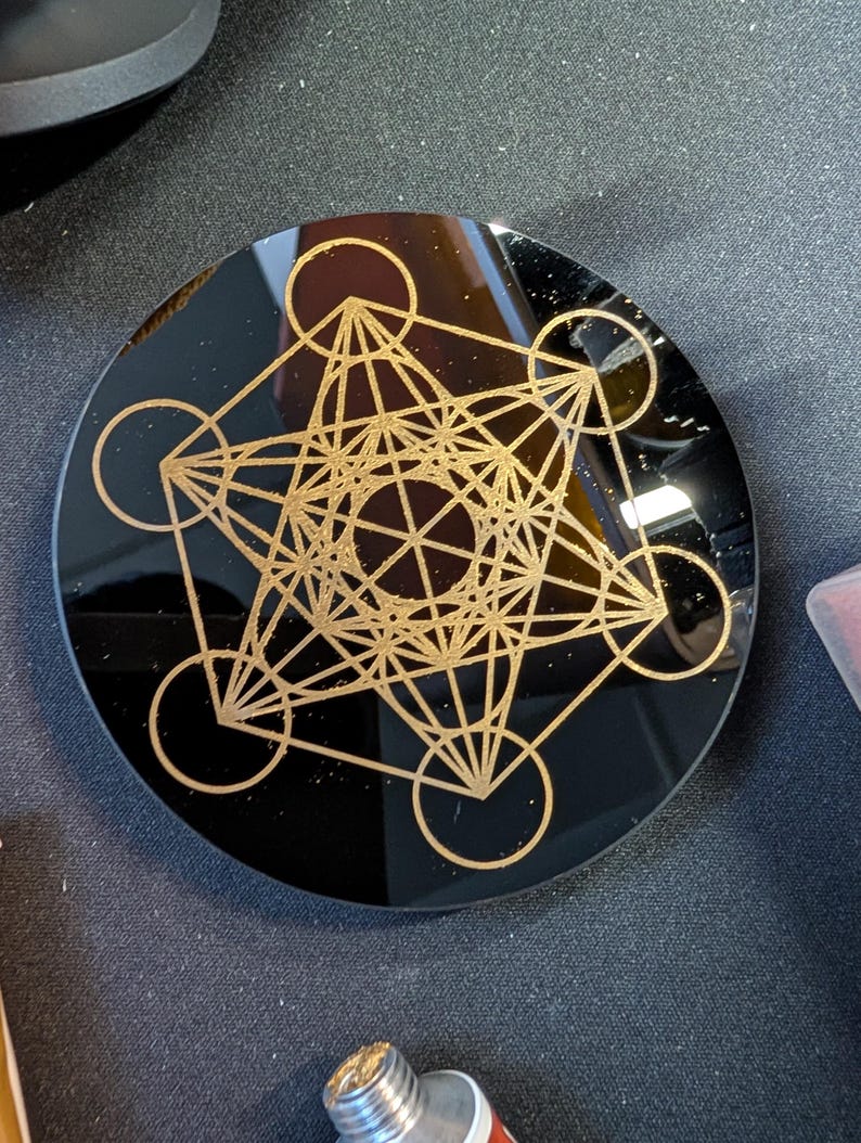 May include: A circular black coaster featuring a gold-colored Metatron's Cube geometric design etched onto the surface. The design consists of interconnected circles and straight lines forming a complex, symmetrical pattern centered on the disc.