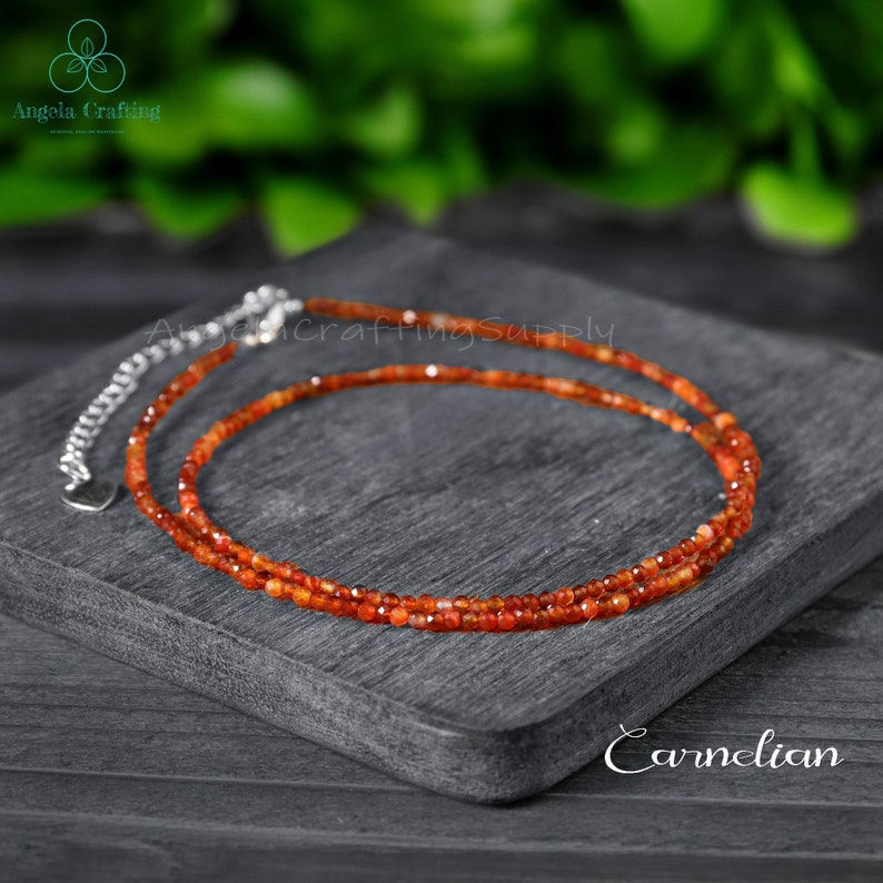 May include: A delicate carnelian necklace with a silver clasp, displayed on a dark gray surface. The necklace is composed of small, faceted orange-red beads. The word "Carnelian" is written in white script.
