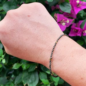 May include: A delicate bracelet featuring small, faceted dark grey beads arranged in a continuous strand, accented by three evenly spaced, smooth, circular gold-toned metal beads that provide a subtle contrast to the dark, textured bead surface.