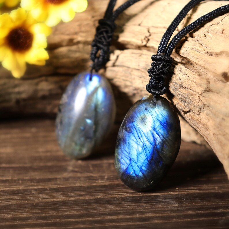 May include: Two oval labradorite pendants with iridescent blue and green hues, suspended on black cords. The gemstones are smooth and polished, showcasing natural patterns. The background includes wood and yellow flowers.