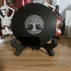 May include: A circular black plaque featuring a silver-toned engraved tree of life design. The plaque is supported by a black, two-piece display stand. The tree motif displays intricate branches, leaves, and roots centered on the flat surface.