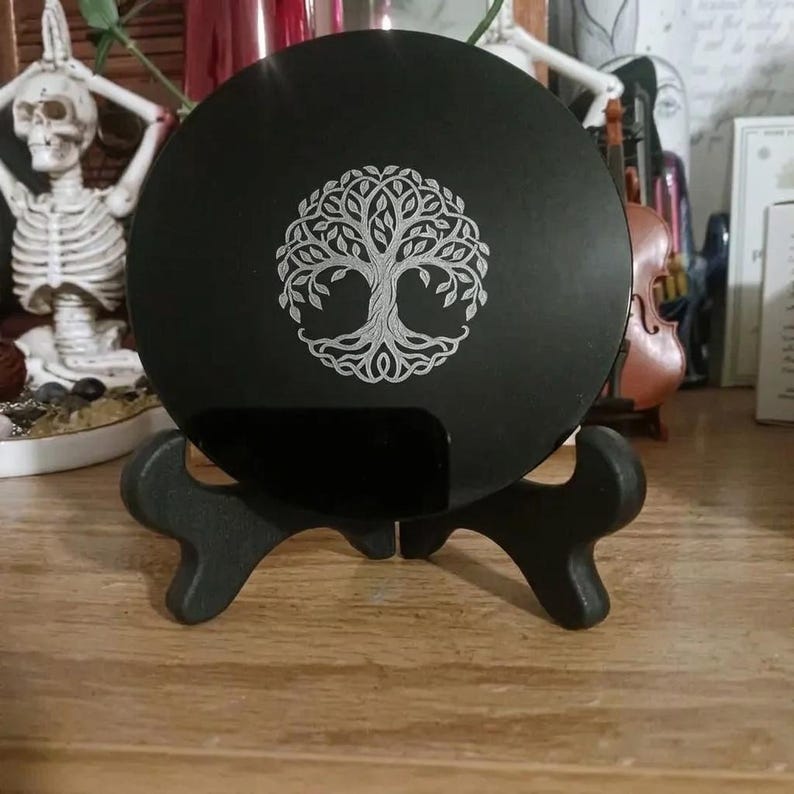 May include: A circular black plaque featuring a silver-toned engraved tree of life design. The plaque is supported by a black, two-piece display stand. The tree motif displays intricate branches, leaves, and roots centered on the flat surface.