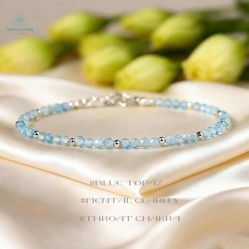 May include: A delicate bracelet featuring small, faceted light blue beads interspersed with smooth, round silver-toned metal beads. The text #BLUE TOPAZ, #MENTAL CLARITY, and #THROAT CHAKRA is visible below the jewelry piece on the fabric surface.