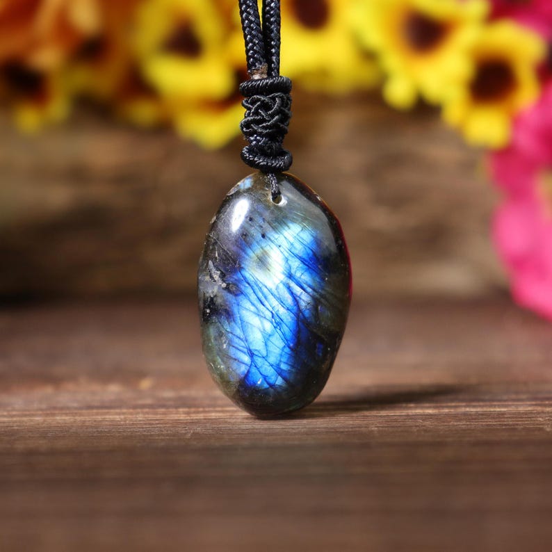 May include: A polished, oval-shaped labradorite pendant with a black cord. The gemstone displays iridescent blue and gray hues. The pendant is suspended from a knotted cord, showcasing a natural stone necklace.