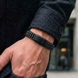 May include: A black bracelet featuring three rows of small, square-shaped beads and a smooth, rectangular matte black clasp. The bracelet is worn on a wrist, partially covered by the sleeve of a dark grey, textured wool-blend jacket.