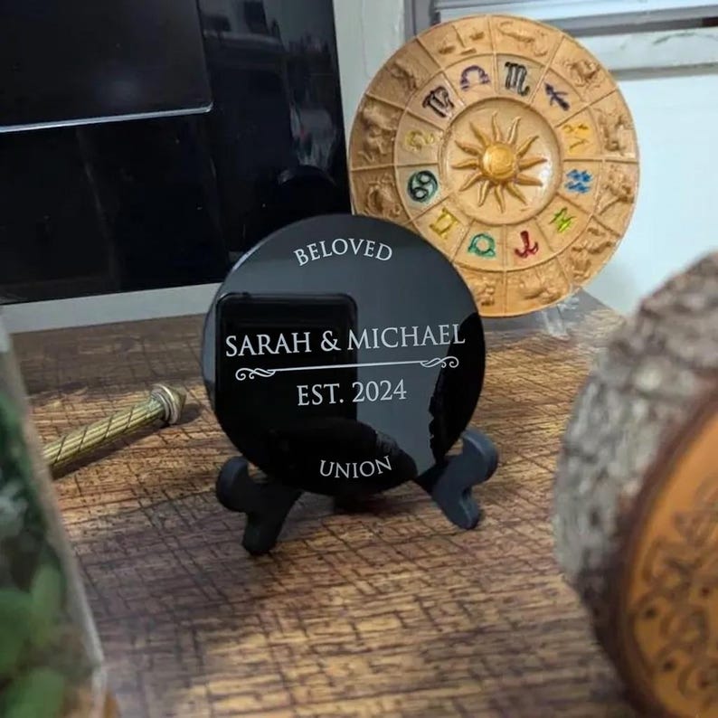 May include: A circular black acrylic plaque displayed on a black stand, featuring white engraved text that reads BELOVED, SARAH & MICHAEL, EST. 2024, and UNION. The plaque is positioned near a circular tan zodiac wheel and a wooden object.