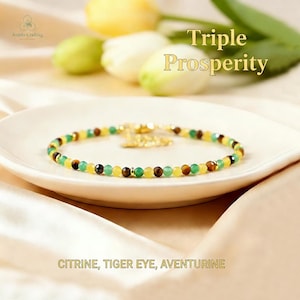 May include: A beaded bracelet with citrine, tiger eye, and aventurine gemstones. The bracelet is on a white plate, with text that reads "Triple Prosperity" and "Citrine, Tiger Eye, Aventurine". The bracelet has gold accents and a gold clasp.
