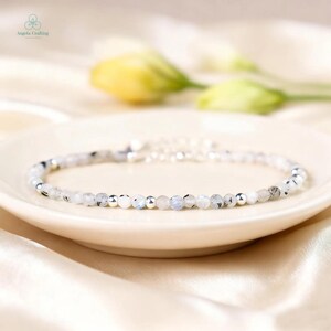 May include: A delicate bracelet featuring small, round, white and gray beads interspersed with silver-colored beads. The bracelet is displayed on a white plate, with soft, blurred flowers in the background. The bracelet is a jewelry item.