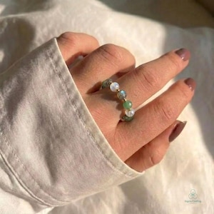 May include: A beaded ring featuring a sequence of small, round, multi-colored gemstones including white, grey, and green beads, interspersed with tiny gold-toned metal spacers, arranged in a circular band design on a finger.