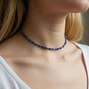 Puede incluir: Un delicado collar con pequeñas cuentas azul oscuro. El collar se lleva alrededor del cuello, mostrando su diseño simple pero elegante. Las cuentas parecen estar espaciadas uniformemente, creando un brillo sutil.