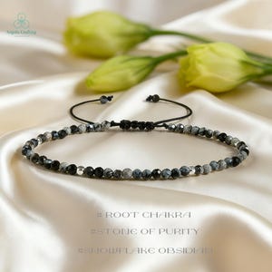 Peut inclure: Un bracelet de perles noires et grises avec une fermeture à nœud coulissant. Le bracelet est composé de petites perles rondes et est présenté sur une surface en satin crème. Le texte de l'image indique "#ROOT CHAKRA", "#STONE OF PURITY", et "#SNOWFLAKE OBSIDIAN".