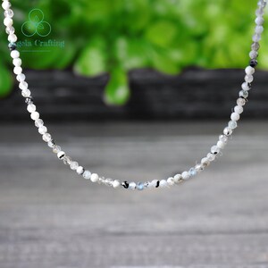 May include: Delicate beaded necklace featuring small, round, white and gray beads. The necklace has a subtle sparkle and a delicate appearance. The beads are strung together, creating a simple yet elegant accessory. The background is blurred, with a green and gray color scheme.