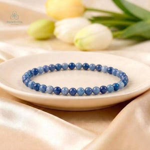 May include: A circular beaded bracelet composed of small, smooth, spherical stones in varying shades of blue, ranging from light sky blue to deep navy, arranged in a continuous loop on a white ceramic plate.