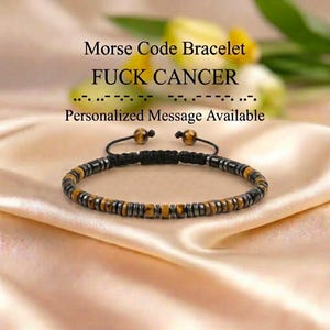 Pode incluir: Uma pulseira de código Morse com contas alternadas de olho de tigre castanho e metal cinzento. A pulseira tem um cordão preto com um fecho de nó deslizante ajustável. O texto acima diz: Morse Code Bracelet FUCK CANCER com pontos e traços.