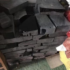 May include: A stack of rectangular dark gray stone blocks with rough, textured surfaces. The stones are arranged in multiple layers, showing varying thicknesses and irregular edges. Some blocks feature smooth, dark, reflective patches on their tops.