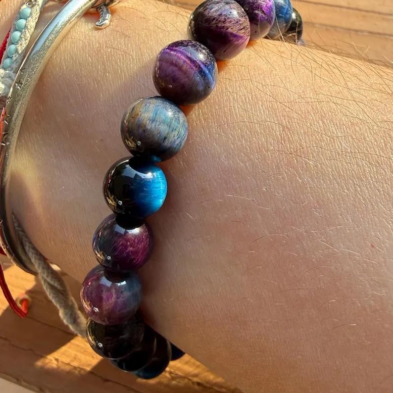 May include: A beaded bracelet featuring smooth, spherical stones in shades of deep purple, vibrant blue, and dark charcoal. The beads exhibit a chatoyant, shimmering luster across their surfaces, arranged in a single strand around the wrist.