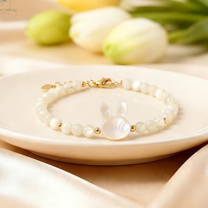 May include: A beaded bracelet featuring a central white mother-of-pearl rabbit charm, accented with small round white beads and gold-toned metal spacers. The bracelet includes a gold-toned lobster claw clasp and a small heart-shaped metal charm.