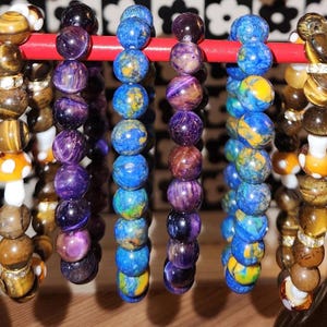 May include: A collection of five beaded bracelets displayed on a red horizontal bar. The bracelets feature round beads in various colors including brown tiger eye, deep purple, vibrant blue, and mixed blue and yellow patterns, with some white mushroom accents.