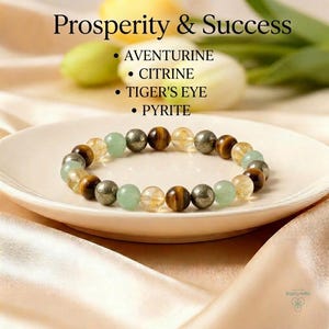 May include: A beaded bracelet featuring aventurine, citrine, tiger's eye, and pyrite gemstones. The bracelet is displayed on a white plate with a soft, neutral background. The text "Prosperity & Success" is at the top.