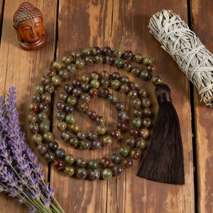 May include: A green and brown beaded mala necklace with a dark brown tassel, a small wooden Buddha statue, a bundle of sage, and a bouquet of lavender on a wooden surface. The beads are approximately 8mm in diameter.