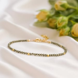 May include: A delicate bracelet featuring small, metallic, dark grey spherical beads interspersed with tiny, polished gold-toned square spacers. The bracelet includes a gold-toned metal clasp mechanism at the end for secure fastening.