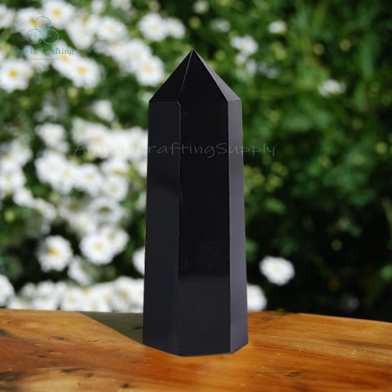 May include: A polished black obsidian crystal tower with a hexagonal base and a pointed top. The stone has a smooth, reflective surface and stands upright, displaying a solid, opaque black color throughout its entire geometric structure.