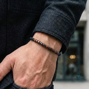 May include: A beaded bracelet featuring alternating cylindrical dark brown wooden beads and small metallic grey beads. The bracelet is worn on a wrist, partially covered by the sleeve of a dark grey textured jacket, showcasing a simple design.