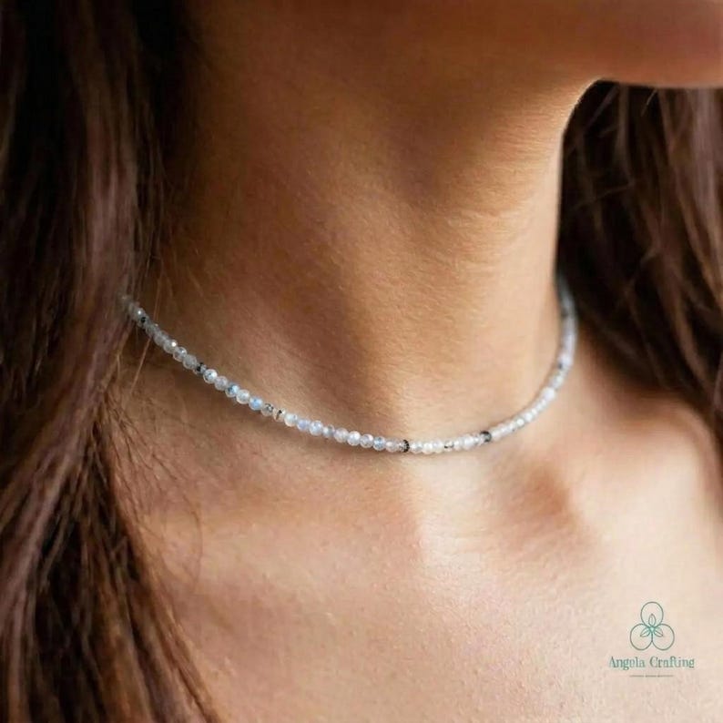 May include: A delicate beaded necklace with small, round, light-colored beads. The necklace is worn around the neck and appears to be made of natural stones or crystals. The beads have subtle variations in color, with some darker accents.