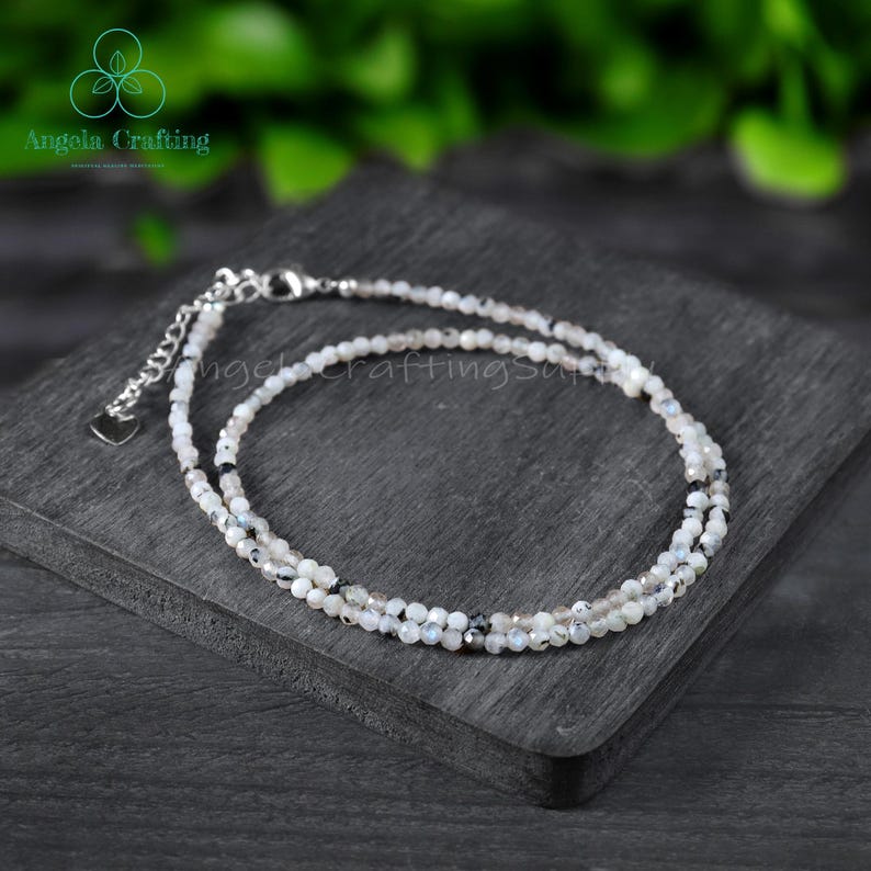 May include: A delicate, layered bracelet featuring small, round beads in shades of white, gray, and black. The bracelet has a silver-toned chain and clasp, with a small heart charm. The jewelry is displayed on a dark gray surface.