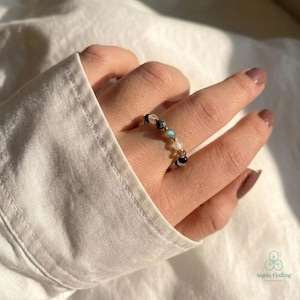 May include: A beaded ring featuring a sequence of small, round gemstones in varying colors, including black, translucent white, iridescent blue, and clear beads, strung together on a thin, flexible band that encircles the finger.