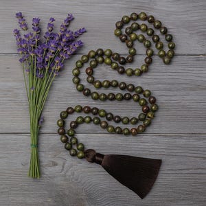 May include: A green and brown beaded mala necklace with a brown tassel, next to a bundle of purple lavender. The beads are round and appear to be made of stone. The lavender is tied with green twine.