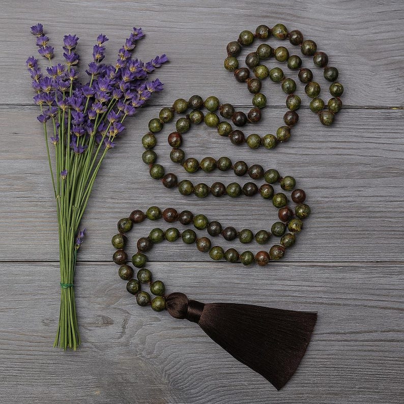 May include: A green and brown beaded mala necklace with a brown tassel, next to a bundle of purple lavender. The beads are round and appear to be made of stone. The lavender is tied with green twine.